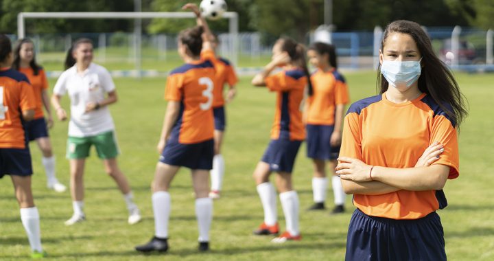 A girl at soccer practice wearing a face mask on the sidelines.